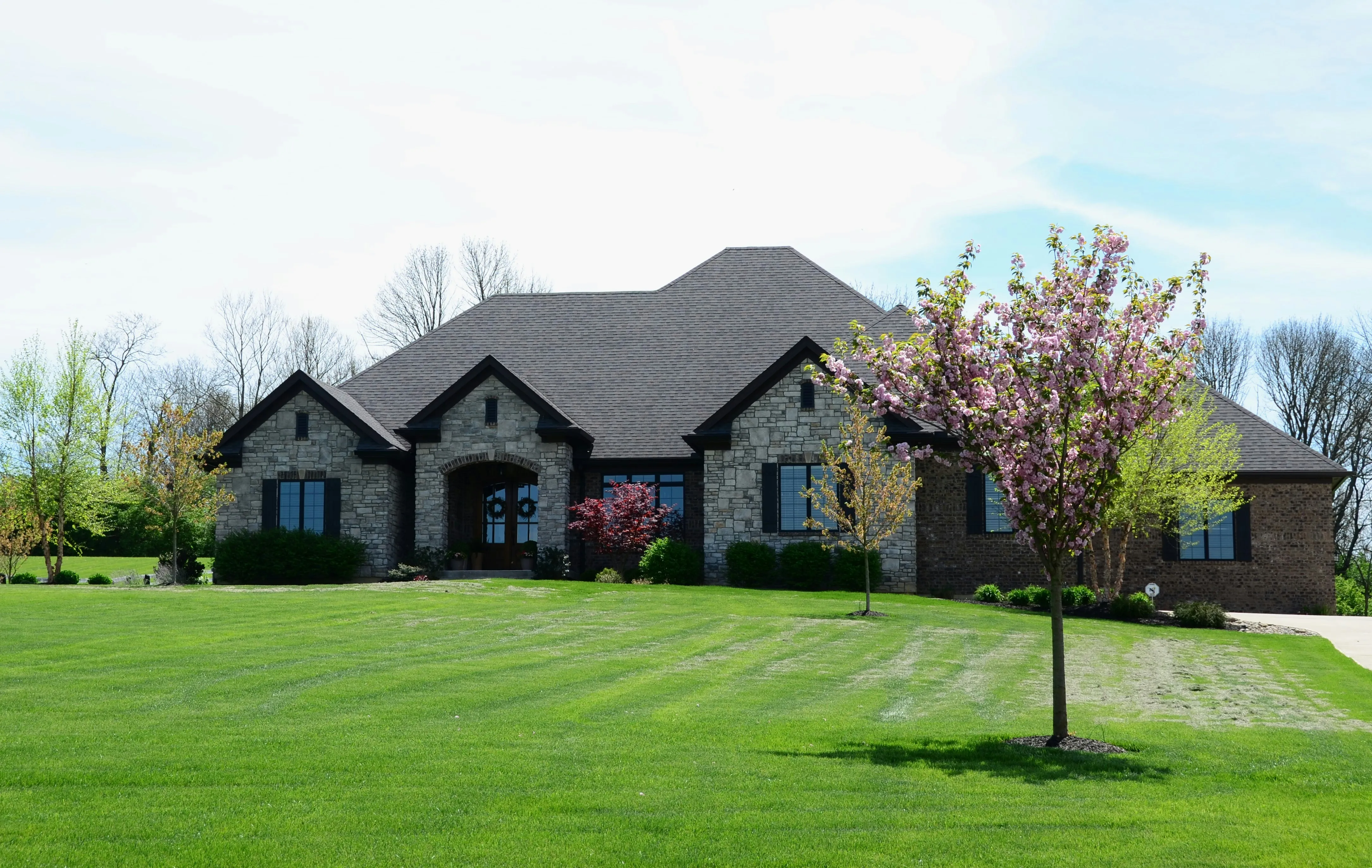 Large stone and brick ranch home with new dark charcoal hip roof surrounded by manicured green lawn, completed residential roofing project